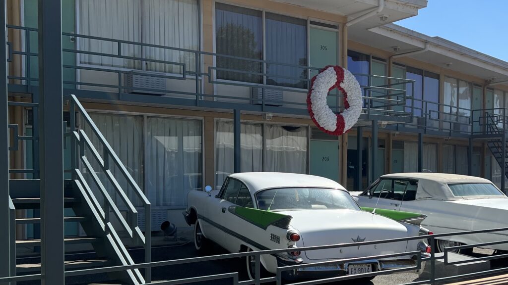 A photo of the Lorraine Hotel. There is a flower wreath at the spot of MLK's death. The hotel has been preserved to what it looked like then with two cars from the time parked outside.